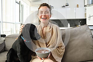 Home and lifestyle concept. Beautiful young woman with dog, sitting on sofa, eating cereals and hugging her puppy, girl