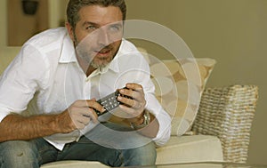 Home lifestyle portrait of young excited and attractive man in white shirt and jeans watching television sitting on living room