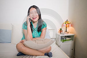 Home lifestyle portrait of young happy and attractive Asian Korean woman sitting on bed talking on mobile phone with girlfriend