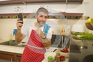 Home lifestyle portrait of young happy and attractive home cook man in red apron following online recipe on internet mobile phone