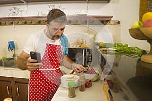Home lifestyle portrait of young happy and attractive home cook man in red apron following online recipe on internet mobile phone