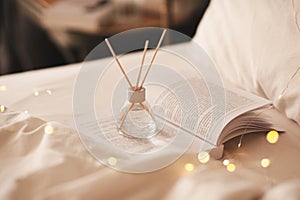 Home liquid fragrance in glass bottle with bamboo sticks stay on open paper book in bed with glow Christmas lights close up.