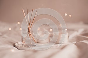 Home liquid fragrance in glass bottle and burning candles staying on white ceramic tray in bed close up.