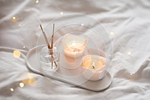 Home liquid fragrance in glass bottle and burning candles staying on white ceramic tray in bed close up.