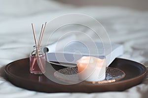 Home liquid fragrance in glass bottle with wooden sticks and scent candle with open paper book on tray in bed