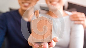 Home loan and financial concept - Close up wooden home toy in hand with blurred asian couple smiling for home loan success