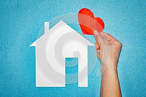 Home love concept. female hand with red heart over white paper house on blue background