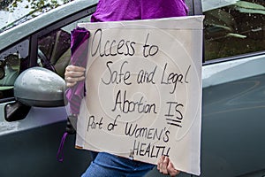 Home made sign reading Access to Safe and Legal Abortion Is part of womens health held by cropped woman in purple shirt holding photo