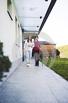 Home nurse walking along a senior woman giving her support to the entrance of her house, vertical photo