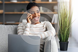 Home Office. Black Freelancer Woman Using Cellphone And Laptop In Living Room.