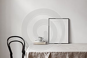 Home office concept. Empty black vertical picture frame mockup. Cup of coffee on old books, linen table cloth. White