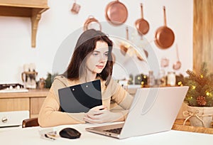 Home office concept. Young woman freelancer sits at a table at home in a bright kitchen in winter, using a laptop and smartphone.