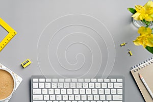 Home office desk with computer keyboard, yellow stationery, flowers, coffee cup and notebook on grey background. Flat lay, top