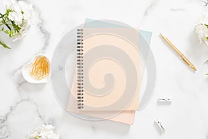 Home office desk table with pastel beige notebook, golden stationery and white flowers on marble background. Flat lay, top view.