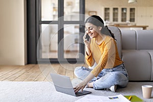 Home Office. Smiling Young Indian Woman Using Laptop And Talking On Cellphone