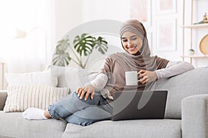 Home Passtime . Cheerful Arabic Woman Watching Movies On Laptop And Drinking Coffee