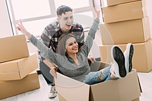 Home, people, moving and real estate concept - happy couple having fun and riding in cardboard boxes at new home