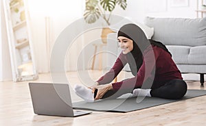 Home Pilates. Sporty arabic woman stretching leg muscles in front of laptop