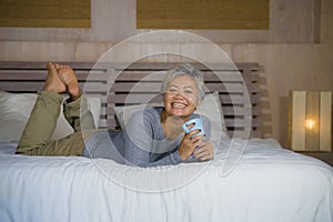 Home portrait of attractive and successful mature Asian American woman with grey hair sitting on bed drinking coffee relaxed