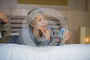 Home portrait of attractive and successful mature Asian American woman with grey hair sitting on bed drinking coffee relaxed