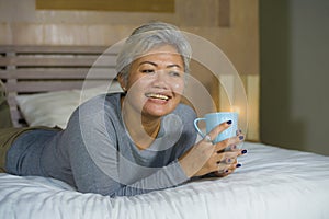 Home portrait of attractive and successful mature Asian American woman with grey hair sitting on bed drinking coffee relaxed