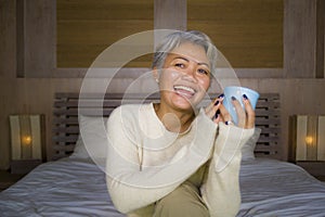 Home portrait of attractive and successful mature Asian American woman with grey hair sitting on bed drinking coffee relaxed