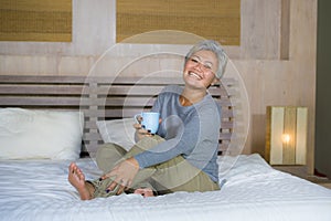 Home portrait of attractive and successful mature Asian American woman with grey hair sitting on bed drinking coffee relaxed