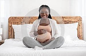 Home Portrait Of Beautiful Pregnant African Woman Sitting On Bed Embracing Belly