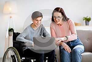 Home schooling concept. Disabled teen boy in wheelchair studying new online materials together with his mother indoors