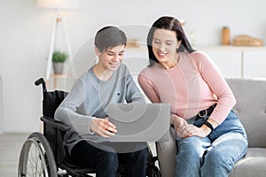 Home schooling. Handicapped teen boy in wheelchair learning online together with his mother indoors