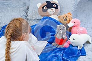 Home schooling. Schoolgirl reading textbook on the bed. Baby girl reading a story to toys