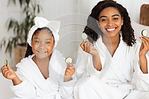 Home Spa. Beautiful African Mother And Daughter In Bathrobes Holding Cucumber Slices