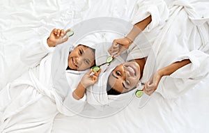 Home Spa. Happy African Mom And Daughter In Bathrobes With Cucumber Slices