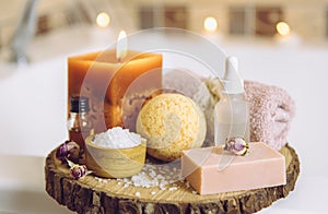 Home spa products on wooden disc tray: bar of soap, bath bomb, aroma bath salt, essential and massage oils, candle burning, rolled