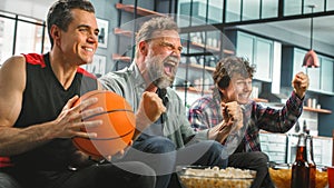 At Home Three Happy Basketball Fans Sitting on a Couch Watch Game on TV, Celebrate Scoring, their