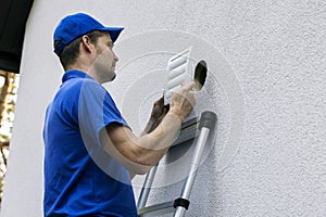 Home ventilation - hvac worker standing on ladder and installing air duct grille on house facade