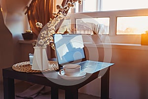 Home work place with laptop, cup of hot drink and blooming brunch in vase on coffee table near window on sunset or sunrise.