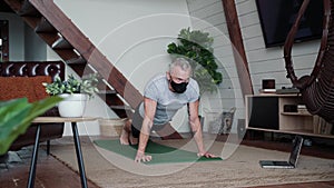 Home workout online. Strong athletic man wearing protective mask doing push ups on yoga mat while exercising at home