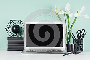 Home workspace with blank laptop monitor, black stationery, books, modern sculpture, coffee cup, white flowers in green mint.
