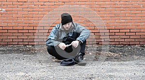 Homeless beggar holds cash money in hands. Refugee needs help. Tramp sits at brick wall wearing in dirty clothes, hat cap. Below