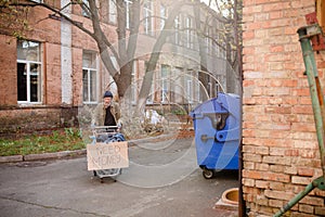 Homeless beggar with stolen shopping cart with board need money on it.