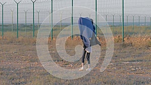 Homeless child walks near protected area. Skinny refugee girl head covered quilt goes along the iron fence state border. Concept: