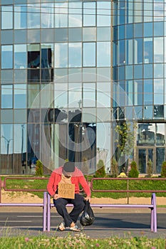 A homeless guy sits on the bench with a cardboard and an inscription: need money. In the background is a business center