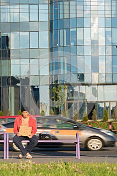 A homeless guy sits on the bench with a cardboard and an inscription: need money. In the background is a business center