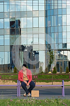 A homeless guy sits on the bench with a cardboard and an inscription: need money. In the background is a business center