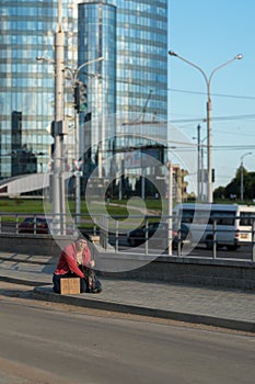 A homeless guy sits on the sidewalk with a cardboard and an inscription: need job. In the background is a business center