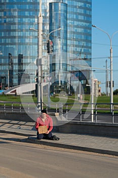 A homeless guy sits on the sidewalk with a cardboard and an inscription: need money. In the background is a business center