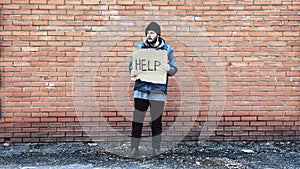Homeless holds piece of cardboard with inscription Help. Homeless is at brick wall looking for job. Male tramp in dirty clothes.