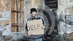 Homeless holds piece of cardboard with inscription Help. Refugee at abandoned building looking for job. Male tramp in dirty