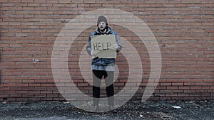 Homeless holds piece of cardboard with inscription Help. Refugee is at brick wall looking for job. Male tramp in dirty